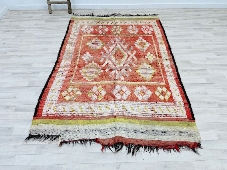 Puede incluir: Una alfombra roja, blanca y verde con un dise&ntilde;o geom&eacute;trico. La alfombra tiene un borde de flecos y est&aacute; hecha de lana.