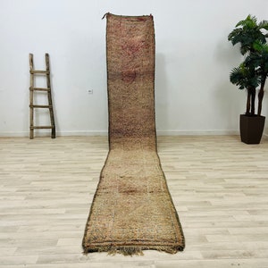 May include: A long, narrow, hand-woven rug with a brown, beige, and pink color scheme. The rug has a fringe on both ends and is laid out on a light wood floor.