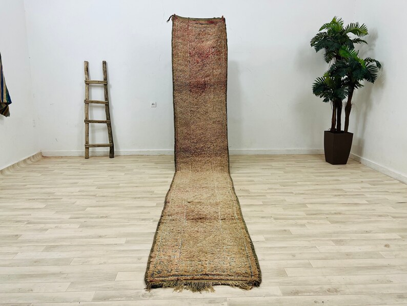 May include: A long, narrow, hand-woven rug with a brown, beige, and pink color scheme. The rug has a fringe on both ends and is laid out on a light wood floor.