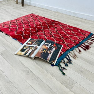 Op de afbeelding: Een rood en roze Marokkaans tapijt met een ruitpatroon en blauwe en witte franjes. Het tapijt ligt op een lichte houten vloer met een tijdschrift erop.