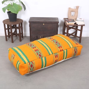 May include: A large, rectangular, orange and green patterned floor cushion with a white underside. The cushion is sitting on a light gray floor in front of a wooden chest and two wooden side tables.