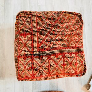 K&ouml;nnte beinhalten: Ein quadratisches Bodenkissen mit einem rot-braun-schwarzen Muster. Das Kissen ist aus gewebtem Stoff und hat ein geometrisches Design.