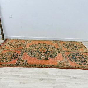 May include: A vintage rug with a geometric pattern in shades of orange, green, and brown. The rug has a fringed edge and is made of wool.