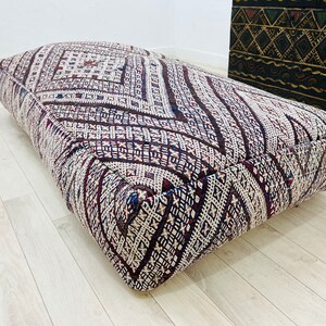 May include: A large, rectangular, patterned floor cushion with a white background and brown, red, and blue geometric designs. The cushion is made of woven fabric and has a thick, comfortable padding.
