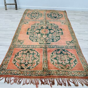 May include: A vintage rug with a traditional Moroccan design. The rug features a repeating pattern of green and cream floral motifs on a peach background. The rug has a fringe border.