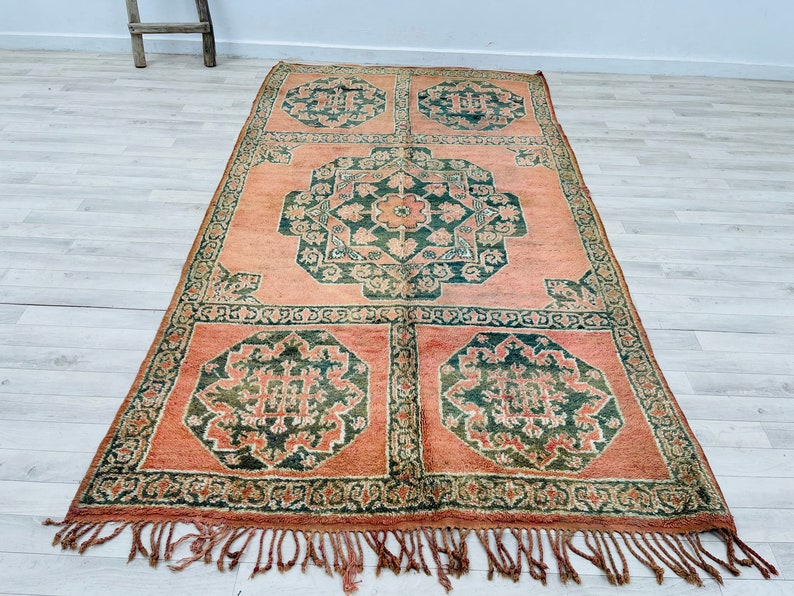May include: A vintage rug with a traditional Moroccan design. The rug features a repeating pattern of green and cream floral motifs on a peach background. The rug has a fringe border.