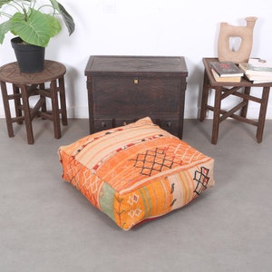 May include: A large, square, orange and beige patterned floor cushion with intricate woven designs. The cushion is sitting on a light gray floor in front of a dark wooden chest with three drawers and two wooden side tables.