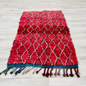 Op de afbeelding: Een rood en zwart Marokkaans tapijt met een ruitpatroon en blauwe franjes. Het tapijt is gemaakt van wol en heeft een zachte, pluche textuur.