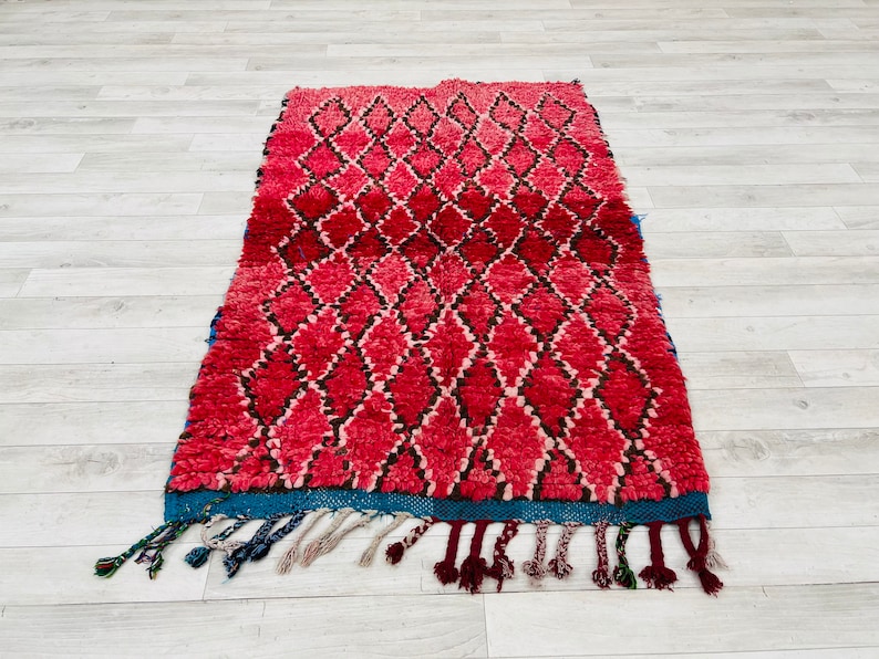 Op de afbeelding: Een rood en zwart Marokkaans tapijt met een ruitpatroon en blauwe franjes. Het tapijt is gemaakt van wol en heeft een zachte, pluche textuur.