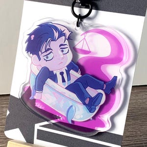 Puede incluir: Un charm de acrílico transparente con un personaje de dibujos animados con cabello oscuro y ropa azul, descansando sobre un diseño rosa y morado. El charm está sujeto a un cierre negro y una tarjeta gris y blanca, exhibido en un pequeño caballete de madera.