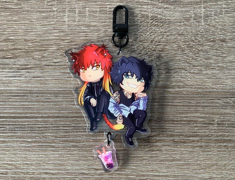 以下が含まれることがあります： 赤と黒の髪の毛をした2人のアニメ風のキャラクターが一緒に座っている、透明なアクリルキーホルダー。キーホルダーには黒い留め金とピンクの飲み物の小さなチャームが付いています。キャラクターは黒と青の衣装を着ています。