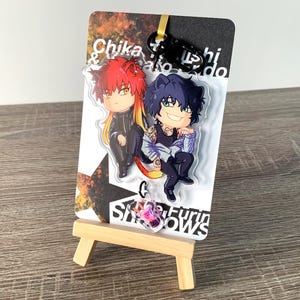 以下が含まれることがあります： 赤と青の髪の毛を持つ2人のアニメスタイルのキャラクターが特徴のアクリルチャーム。チャームは黒いクリップに取り付けられ、テキストと星のデザインが施されたカードに表示されます。カードは小さな木製のイーゼルに立てかけられています。