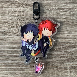 以下が含まれることがあります： 黒と赤の髪の毛をした2人のアニメ風のキャラクターが並んで座っている、透明なアクリルキーホルダー。キーホルダーには、オレンジ色のストローと黒い留め金が付いたピンク色の飲み物の小さなチャームが含まれています。