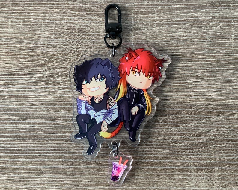 以下が含まれることがあります： 黒と赤の髪の毛をした2人のアニメ風のキャラクターが並んで座っている、透明なアクリルキーホルダー。キーホルダーには、オレンジ色のストローと黒い留め金が付いたピンク色の飲み物の小さなチャームが含まれています。