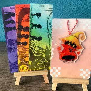 May include: A set of four colorful bookmarks with a silhouette design and a small, red, cartoon character charm on a pink card with a white checkered pattern.