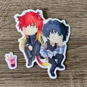 以下が含まれることがあります： 赤い髪と黒い髪の、動物の耳を持つ2人のアニメスタイルのキャラクターが描かれたステッカー。彼らは一緒に座っており、小さな飲み物のステッカーが隣にあります。キャラクターは黒い衣装を着ています。
