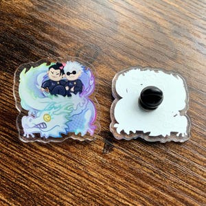 Puede incluir: Un pin de acrílico transparente con dos personajes de dibujos animados montando un dragón azul y blanco. El pin tiene un acabado iridiscente arcoíris y una parte posterior de metal negro.