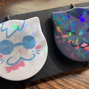 Può includere: Due spille in smalto olografiche iridescenti con volti di animali dei cartoni animati. La prima spilla è bianca con un gatto dagli occhi azzurri che indossa occhiali da sole e un fiocco rosa. La seconda spilla è nera con un gatto dagli occhi verdi con la lingua rosa che sporge.