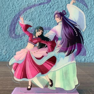 Puede incluir: Un soporte de acrílico transparente con una base morada presenta una ilustración pintada a mano de dos personajes de anime bailando. El personaje femenino lleva un vestido rosa y rojo con una cinta azul. El personaje masculino lleva una túnica blanca y verde con el pelo largo morado.