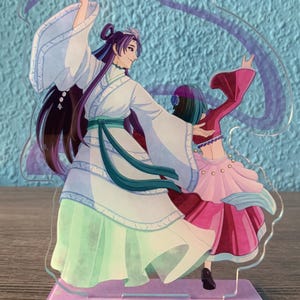 May include: A clear acrylic stand with a colorful illustration of two figures dancing. The figures are wearing elaborate costumes with flowing fabric. The stand has a purple base with a glittery finish.