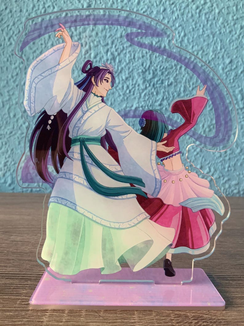 May include: A clear acrylic stand with a colorful illustration of two figures dancing. The figures are wearing elaborate costumes with flowing fabric. The stand has a purple base with a glittery finish.