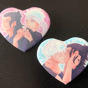 May include: Two heart-shaped pins featuring anime-style characters. The first pin has a pink background with two characters, one with blue hair and the other with black hair. The second pin has a blue background with two characters kissing, one with white hair and the other with black hair.