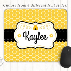 May include: A personalized mousepad with a yellow honeycomb pattern and a black border. The center features a white label with the name "Kaylee" in black script, a small bee hive graphic, and a flower. A black computer mouse is on the right.