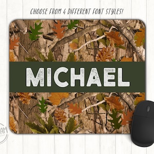 May include: A rectangular mousepad with a camouflage pattern of brown, tan, and green. The name "MICHAEL" is displayed in white block letters on a dark green banner. A black computer mouse is on the right. Text at the top says "CHOOSE FROM 4 DIFFERENT FONT STYLES!"
