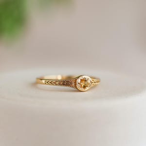 Peut inclure: Une bague en or avec une pierre précieuse jaune ronde sertie dans une lunette détaillée. La bande présente un motif gravé. La bague est présentée sur une surface neutre, soulignant son design complexe et sa couleur chaude.