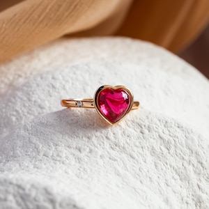 Peut inclure: Une bague en or avec une pierre précieuse rose en forme de cœur et un petit accent diamant. La bague est posée sur une surface blanche.