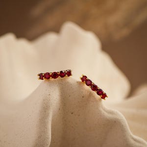 Puede incluir: Dos anillos dorados, cada uno adornado con una fila de gemas rojas vibrantes. Los anillos se presentan sobre un fondo neutro y texturizado, creando un contraste llamativo. Las gemas están espaciadas uniformemente.