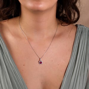 Op de afbeelding: Een zilveren ketting met een hanger met een rode edelsteen omringd door kleinere heldere edelstenen.