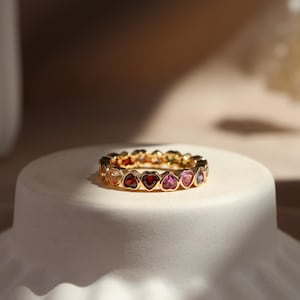 Puede incluir: Un anillo de oro con una serie de piedras preciosas en forma de corazón de varios colores. Las piedras están engastadas juntas, creando una banda completa de color. El anillo se muestra sobre una superficie blanca y redondeada.