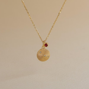 May include: A gold necklace featuring a circular pendant with a textured sunburst finish and a small constellation pattern of tiny embedded stones. A separate, smaller circular charm with a single red stone hangs beside the main pendant.