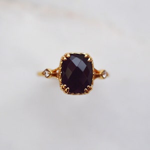 May include: A gold ring featuring a large, faceted, cushion-cut dark purple gemstone held by decorative prongs. The band includes two smaller, clear, round-cut stones set on either side of the central setting, with a textured metal finish.