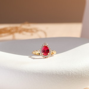 Puede incluir: Un anillo de oro con una piedra preciosa roja en forma de pera en el centro, rodeada por un halo de pequeñas piedras blancas. La banda está hecha de oro e incluye pequeñas piedras blancas adicionales engastadas a lo largo del aro.