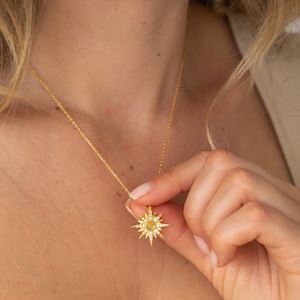 May include: A gold-toned necklace featuring a starburst-shaped pendant. The pendant is adorned with small, clear, sparkling stones radiating outward from a central, circular, light-colored stone, all suspended from a delicate gold-toned chain.