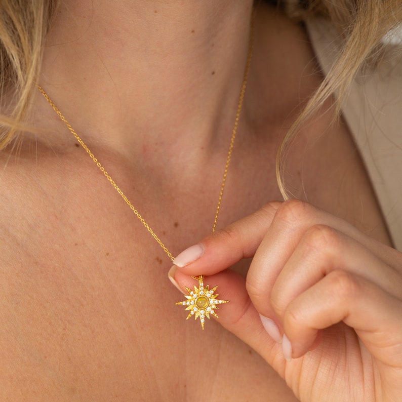 May include: A gold-toned necklace featuring a starburst-shaped pendant. The pendant is adorned with small, clear, sparkling stones radiating outward from a central, circular, light-colored stone, all suspended from a delicate gold-toned chain.
