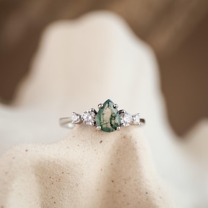 May include: A silver ring featuring a pear-shaped, green moss agate gemstone as the centerpiece, flanked by smaller, clear gemstones. The ring is set on a neutral background, highlighting the jewelry's design and details.