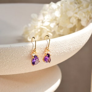 Può includere: Un paio di orecchini pendenti in oro con pietre preziose viola a forma di goccia e piccoli cristalli trasparenti. Gli orecchini sono su una superficie bianca.