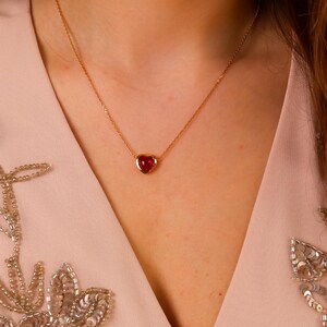 May include: A gold chain necklace featuring a small, smooth, heart-shaped red gemstone pendant set in a gold bezel. The necklace rests against a light pink garment adorned with silver sequin floral embroidery along the neckline edges.