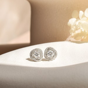 Puede incluir: Un par de pendientes de botón de plata con una piedra redonda y transparente rodeada de piedras más pequeñas en un diseño circular.