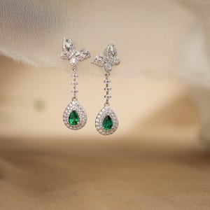 May include: A pair of silver drop earrings. Each earring has a butterfly-shaped top, a series of small, round accents, and a teardrop-shaped pendant with a vibrant green centre stone, surrounded by smaller, clear stones.