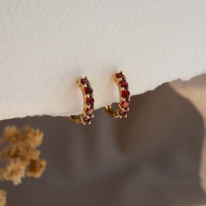 Peut inclure: Une paire de créoles dorées ornées d'une rangée de petites pierres précieuses grenat rondes et rouge foncé, serties le long de la courbe avant de chaque anneau. Les boucles d'oreilles présentent une finition métallique polie et un fermoir à charnière.