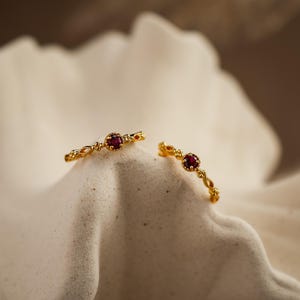 May include: Two delicate gold rings with a small, round, deep red gemstone at the center. The rings have an open design with a series of small, decorative elements. The rings are displayed on a textured, off-white surface.