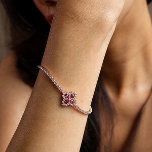 May include: A delicate bracelet featuring a clover-shaped charm set with red rubies and white diamonds. The bracelet is crafted from white gold and has a polished finish.