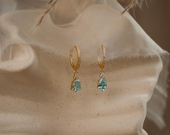 Puede incluir: Un par de pendientes de aro dorados con una pequeña hilera de piedras pavé transparentes a lo largo del aro y un colgante de piedra preciosa azul claro en forma de lágrima, con tres diminutas piedras transparentes en la parte superior.