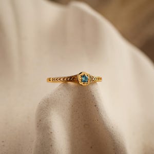 Könnte beinhalten: Ein goldener Ring mit einer kreisförmigen Fassung in der Mitte, die einen kleinen, leuchtend blauen Edelstein enthält. Das Band ist mit einem sich wiederholenden blattartigen Muster verziert, das sich vom zentralen Element aus erstreckt.
