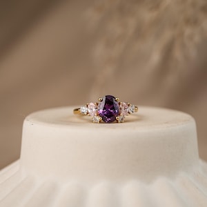Può includere: Un anello in oro con una pietra preziosa centrale di taglio ovale color viola intenso, affiancata da due pietre ovali più piccole color rosa chiaro e quattro minuscole pietre rotonde trasparenti montate su una sottile fascia dorata.