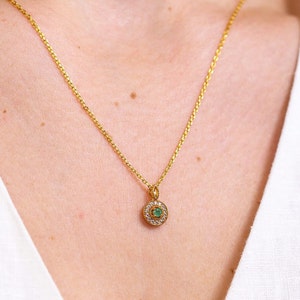 May include: A gold chain necklace featuring a circular pendant with a central round green gemstone surrounded by a halo of small clear white stones. The pendant has a textured gold border and is suspended from a delicate gold link chain.
